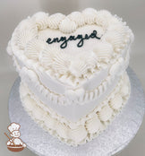 A heart-shaped, single-tier white cake with intricate piped frosting details and the word "engaged" written in black icing on top.