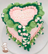 A heart-shaped pink cake with "Happy Birthday" written on top, decorated with green and pink piped frosting, white pearls, and cascading floral designs.