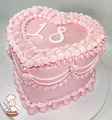 Heart-shaped pink cake with "18" on top, decorated with piped frosting, pearl-like accents, and scalloped edges on a white base.
