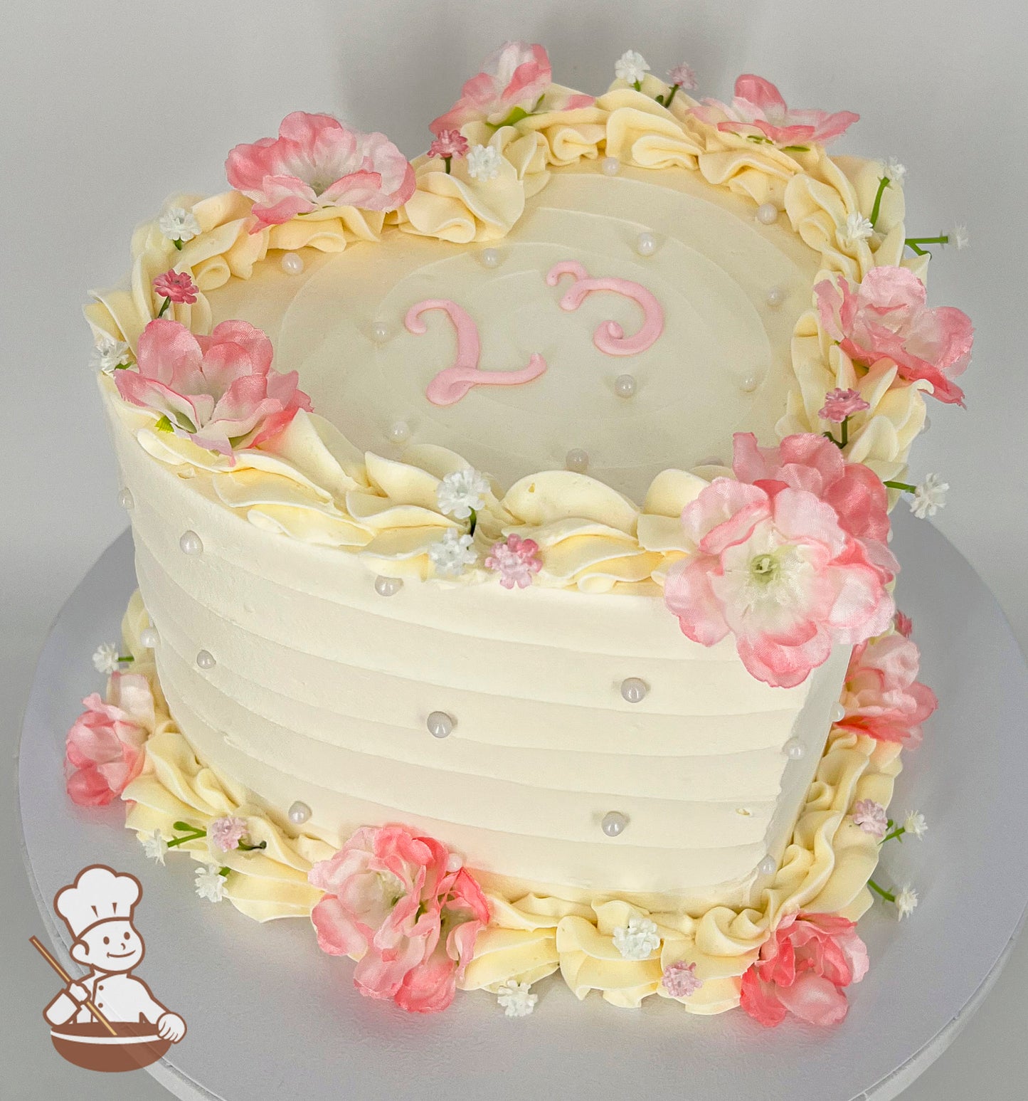 A heart-shaped cake with pastel cream frosting, adorned with pink and white flowers, piped cream ruffles, pearl accents, and "23" in pink icing on top.