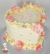 A heart-shaped cake with pastel cream frosting, adorned with pink and white flowers, piped cream ruffles, pearl accents, and "23" in pink icing on top.