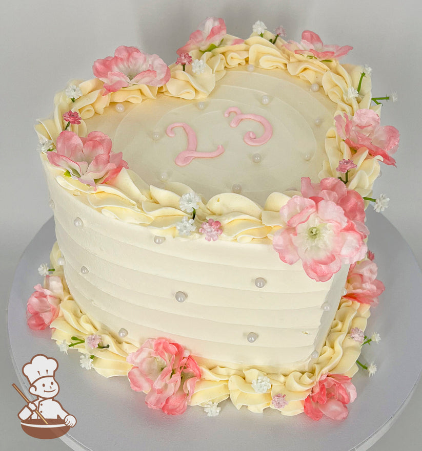 A heart-shaped cake with pastel cream frosting, adorned with pink and white flowers, piped cream ruffles, pearl accents, and "23" in pink icing on top.