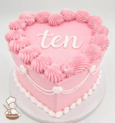 Heart-shaped pink cake with piped frosting swirls along the edges, white decorative piping, and the word "ten" written in white icing on top.