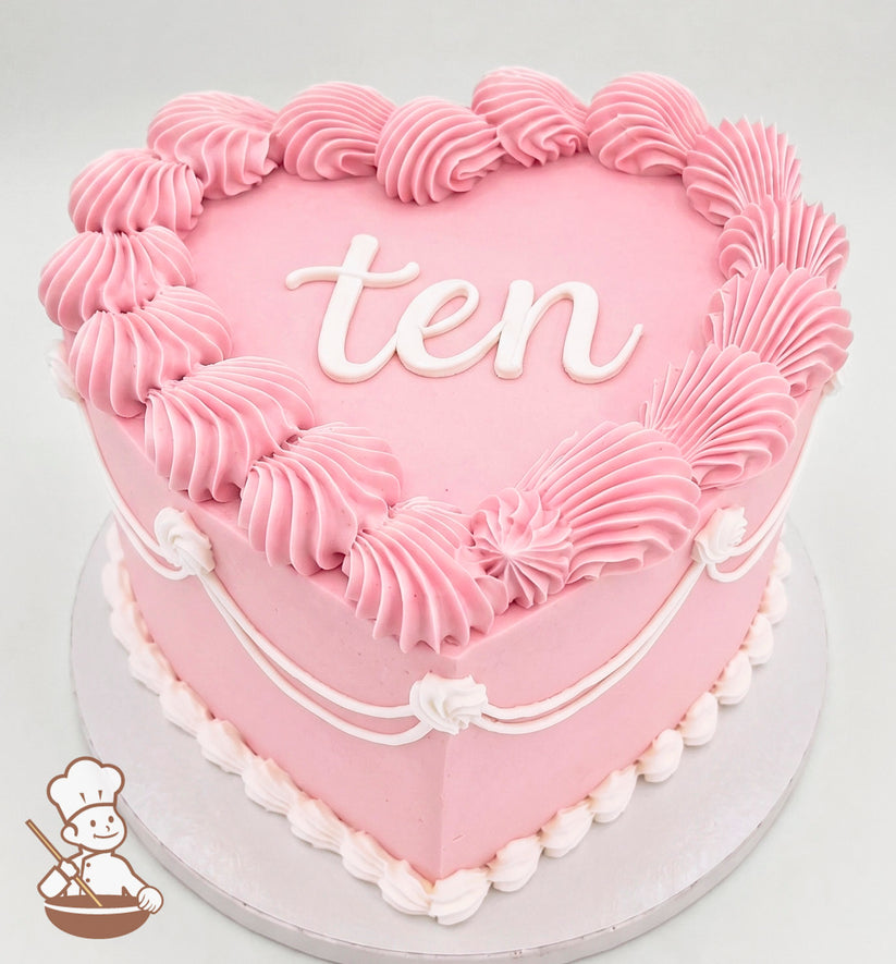 Heart-shaped pink cake with piped frosting swirls along the edges, white decorative piping, and the word "ten" written in white icing on top.