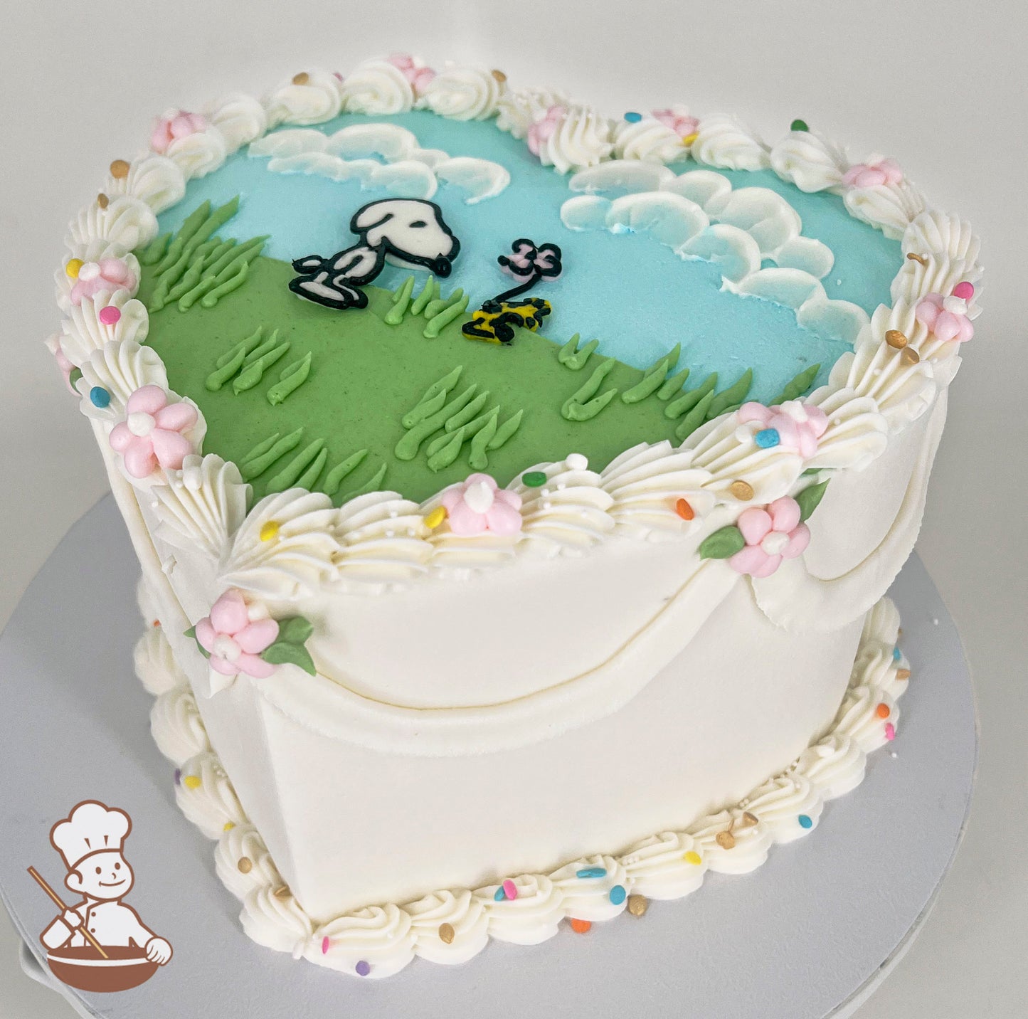 Heart-shaped cake with white frosting, pastel piping, and floral accents. The top features Snoopy and Woodstock on a grassy field with a blue sky and clouds.