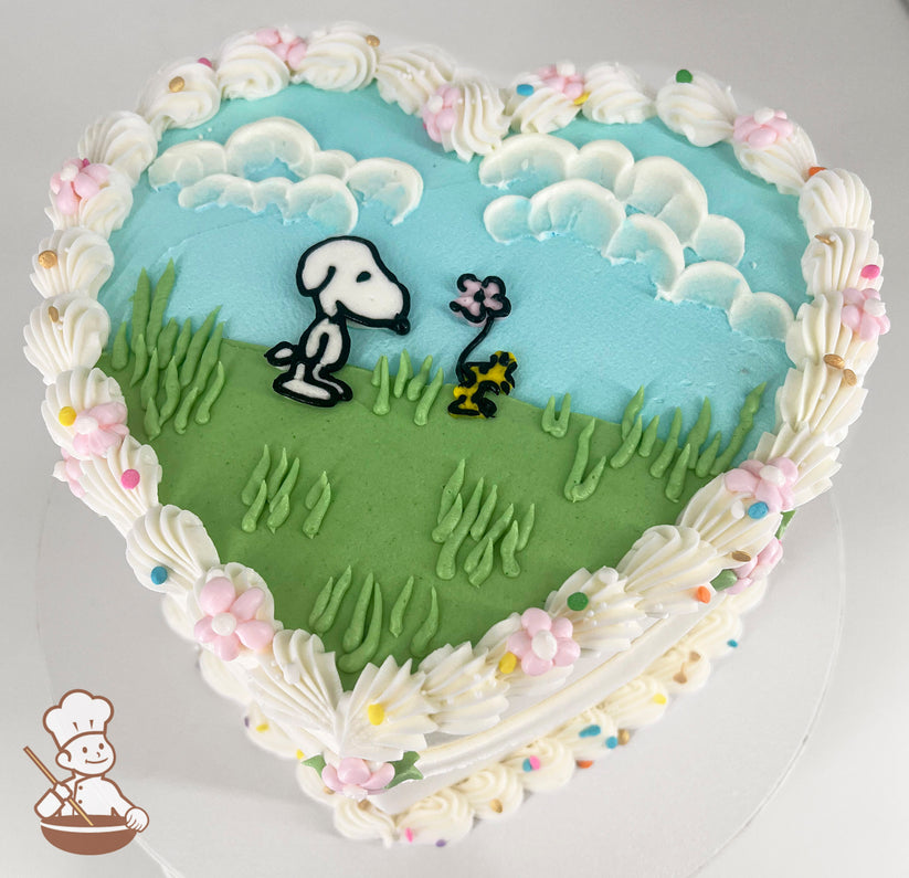 Heart-shaped cake with a pastel blue sky, white clouds, and green grass design. Features a cartoon dog and a small yellow bird near a pink flower. Decorated with piped white frosting, pastel flowers, and sprinkles along the edges.
