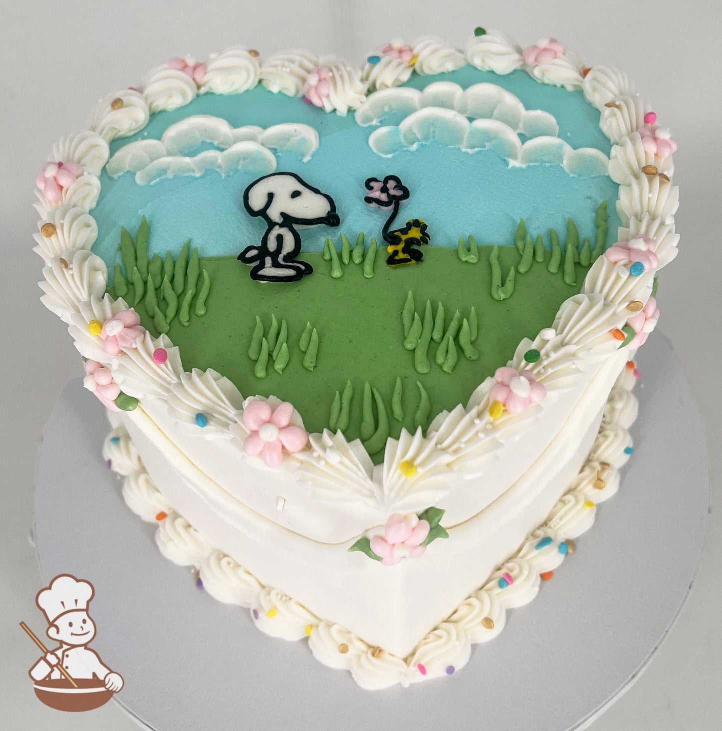 Heart-shaped cake with white frosting, pastel flowers, and colorful sprinkles. Top features Snoopy and Woodstock on green grass under a blue sky with clouds.