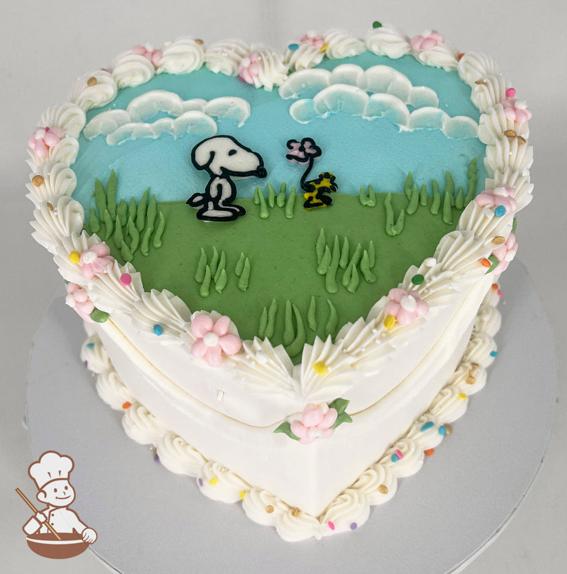 Heart-shaped cake with white frosting, pastel flowers, and colorful sprinkles. Top features Snoopy and Woodstock on green grass under a blue sky with clouds.