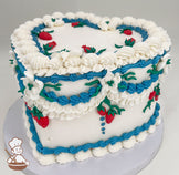 Heart-shaped white cake with piped white and blue frosting, adorned with red strawberry decorations and green leaves. Placed on a white base.