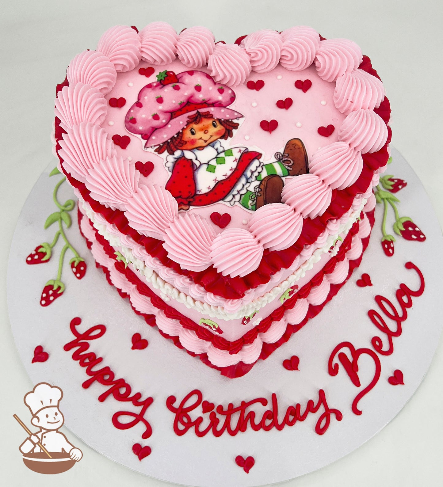 Heart-shaped birthday cake with pink and red frosting, featuring a cartoon character on top. Decorated with small hearts and "Happy Birthday Bella" written on the base.