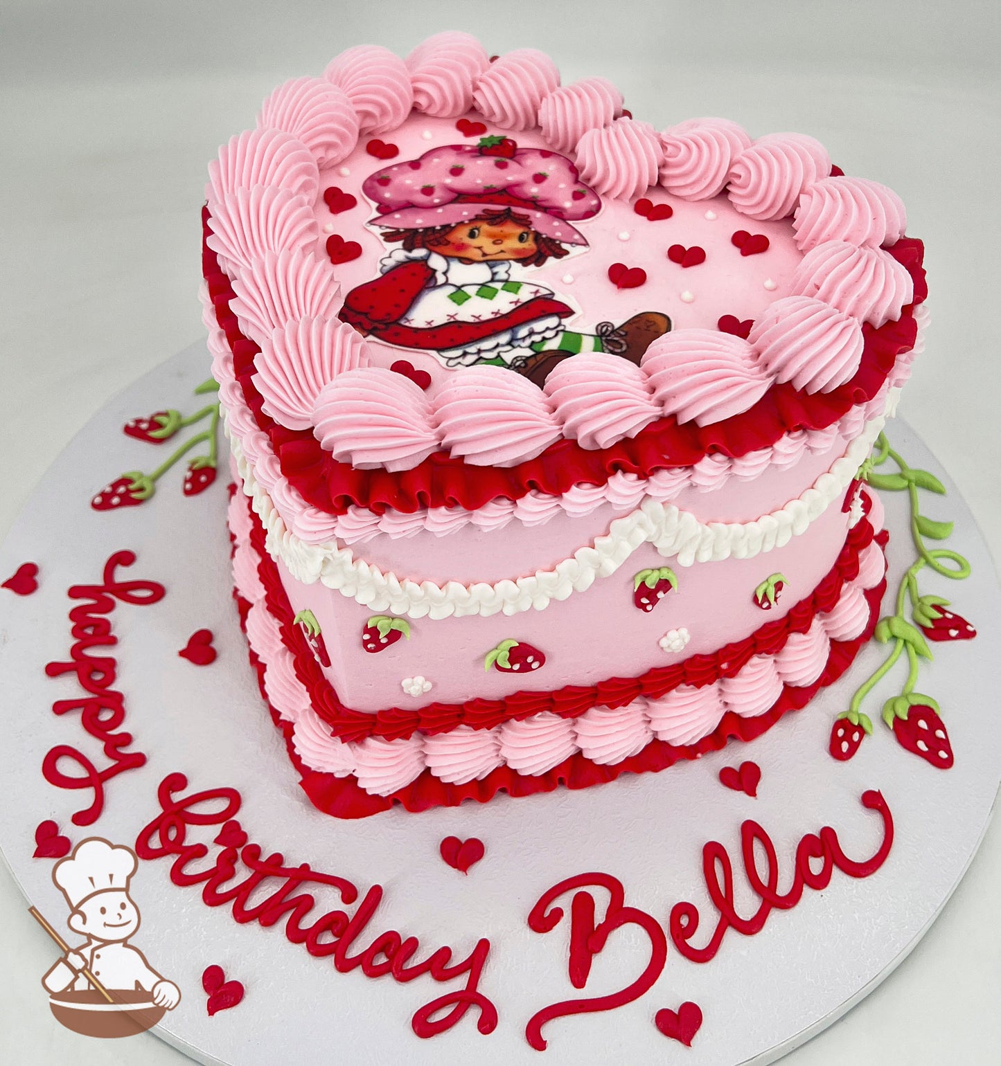 Heart-shaped pink cake with piped pink and red frosting, strawberry decorations, and a cartoon character on top. "Happy Birthday Bella" written on the base.