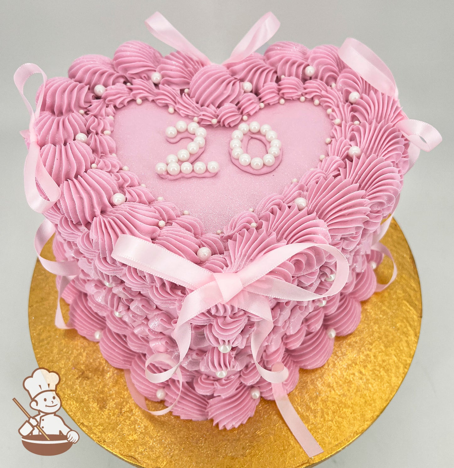 Heart-shaped pink cake with piped frosting ruffles, pearl accents, and pink ribbon bows. The top features the number "20" in white pearls on a glittery surface, placed on a gold board.
