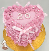 Heart-shaped pink cake with piped frosting ruffles, pearl accents, and pink ribbon bows. The top features the number "20" in white pearls on a glittery surface, placed on a gold board.