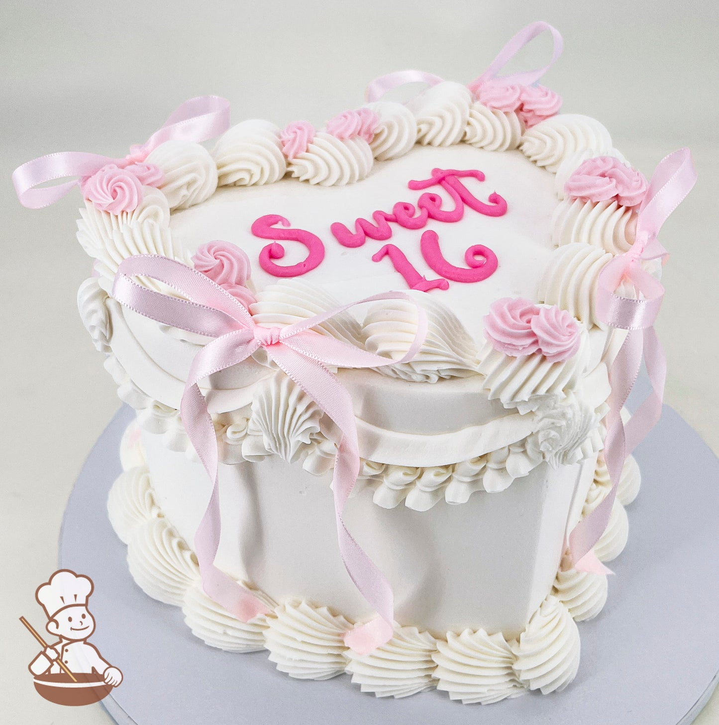 A white, single-tier cake with piped white frosting swirls, pink roses, and pink ribbon bows. "Sweet 16" is written in pink icing on top.
