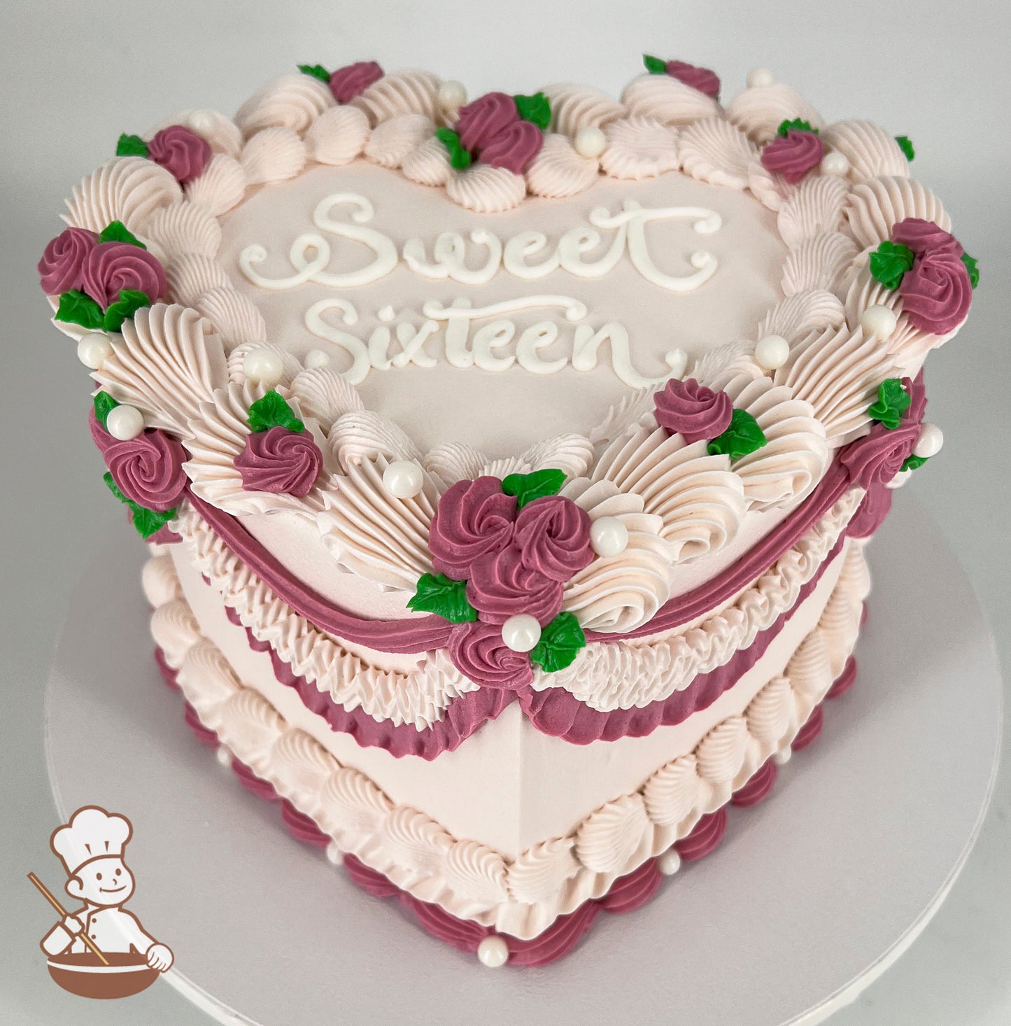 Heart-shaped pink cake with "Sweet Sixteen" written on top, decorated with piped frosting, purple roses, green leaves, and white pearl accents.