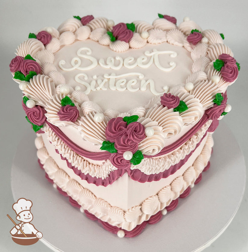 Heart-shaped pink cake with "Sweet Sixteen" written on top, decorated with piped frosting, purple roses, green leaves, and white pearl accents.