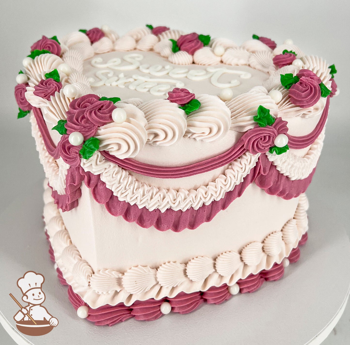 A single-tier cake with pink and white frosting, decorated with piped roses, green leaves, pearl-like accents, and ornate swirls.