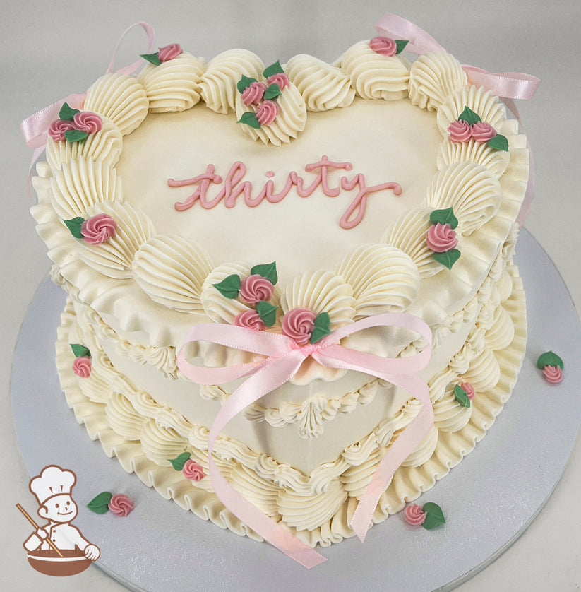 Heart-shaped, single-tier cream-colored cake with piped frosting, pink rose decorations, green leaves, and "thirty" written in pink icing on top.