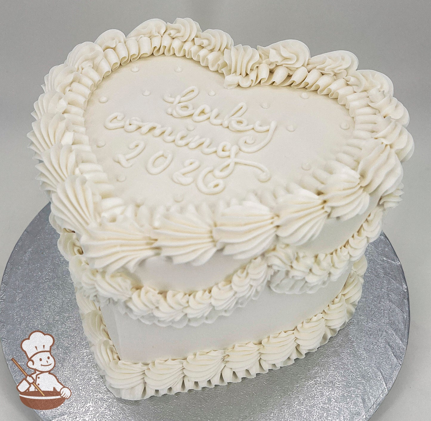 Heart-shaped white cake with piped buttercream ruffles and the text "baby coming 2026" written on top. Displayed on a silver cake board.