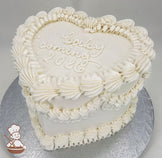 Heart-shaped white cake with piped buttercream ruffles and the text "baby coming 2026" written on top. Displayed on a silver cake board.