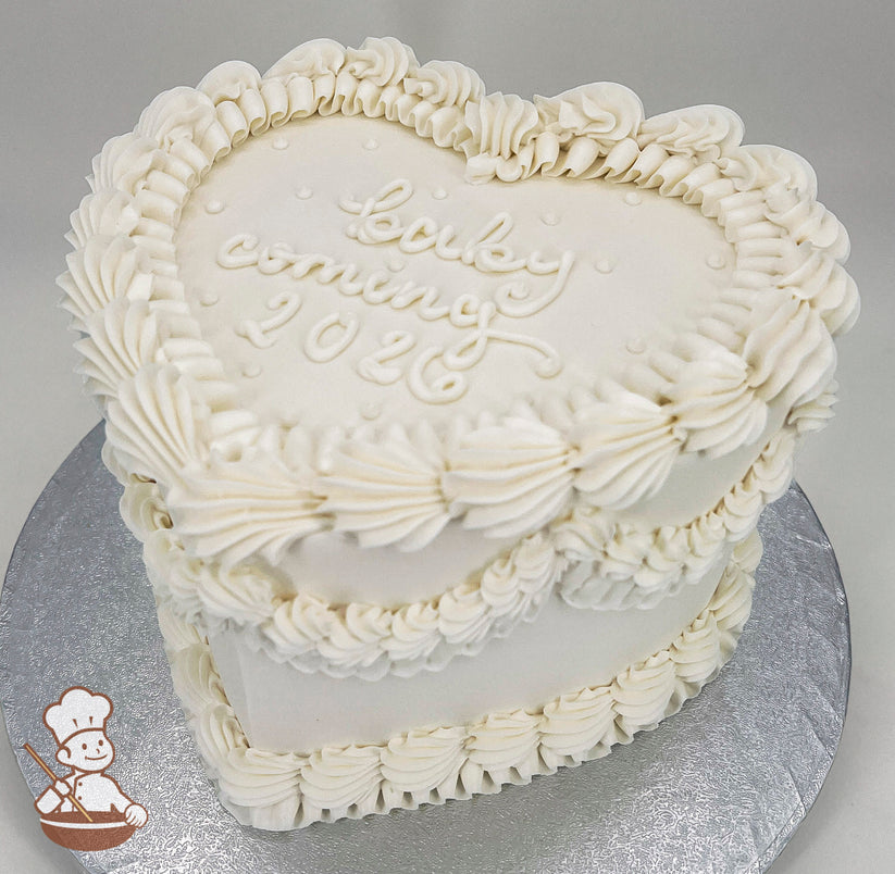 Heart-shaped white cake with piped buttercream ruffles and the text "baby coming 2026" written on top. Displayed on a silver cake board.