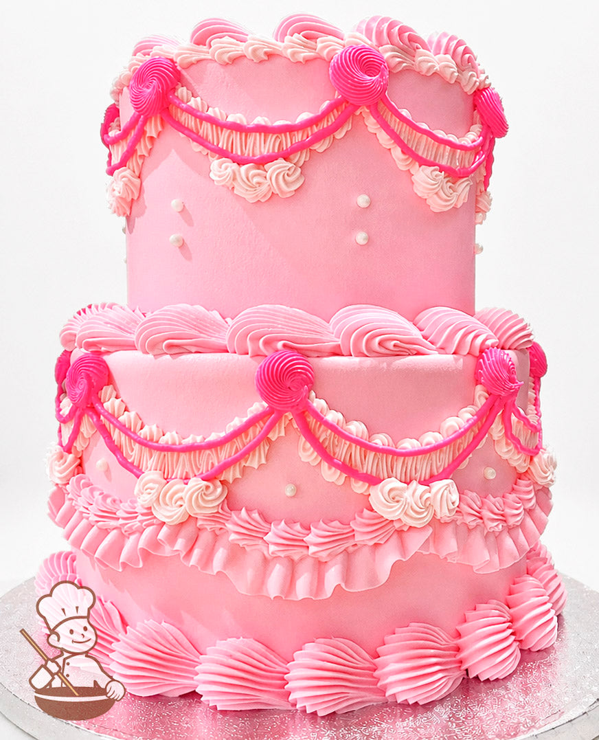 Two-tier pink cake with intricate piping details, including ruffles, swirls, and pearl accents. Decorated with bright pink draped icing and rosettes.