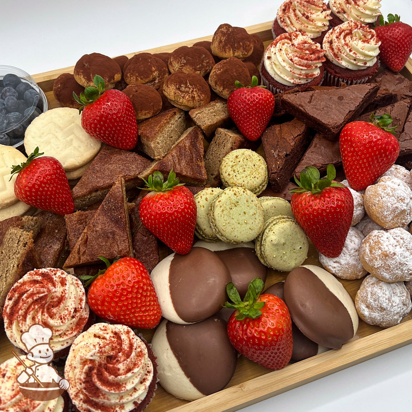 A rectangular bamboo tray carrying red velvet cupcakes, cream puffs, blondies, brownies, shortbreads, a bowl of blueberries, and strawberries.
