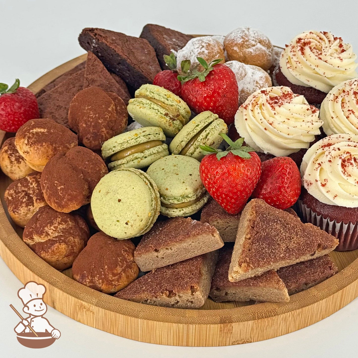 An assortment of red velvet cupcakes, cream puffs, blondies, brownies, pistachio macarons, and strawberries on a round bamboo tray.