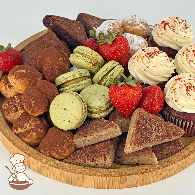 An assortment of red velvet cupcakes, cream puffs, blondies, brownies, pistachio macarons, and strawberries on a round bamboo tray.