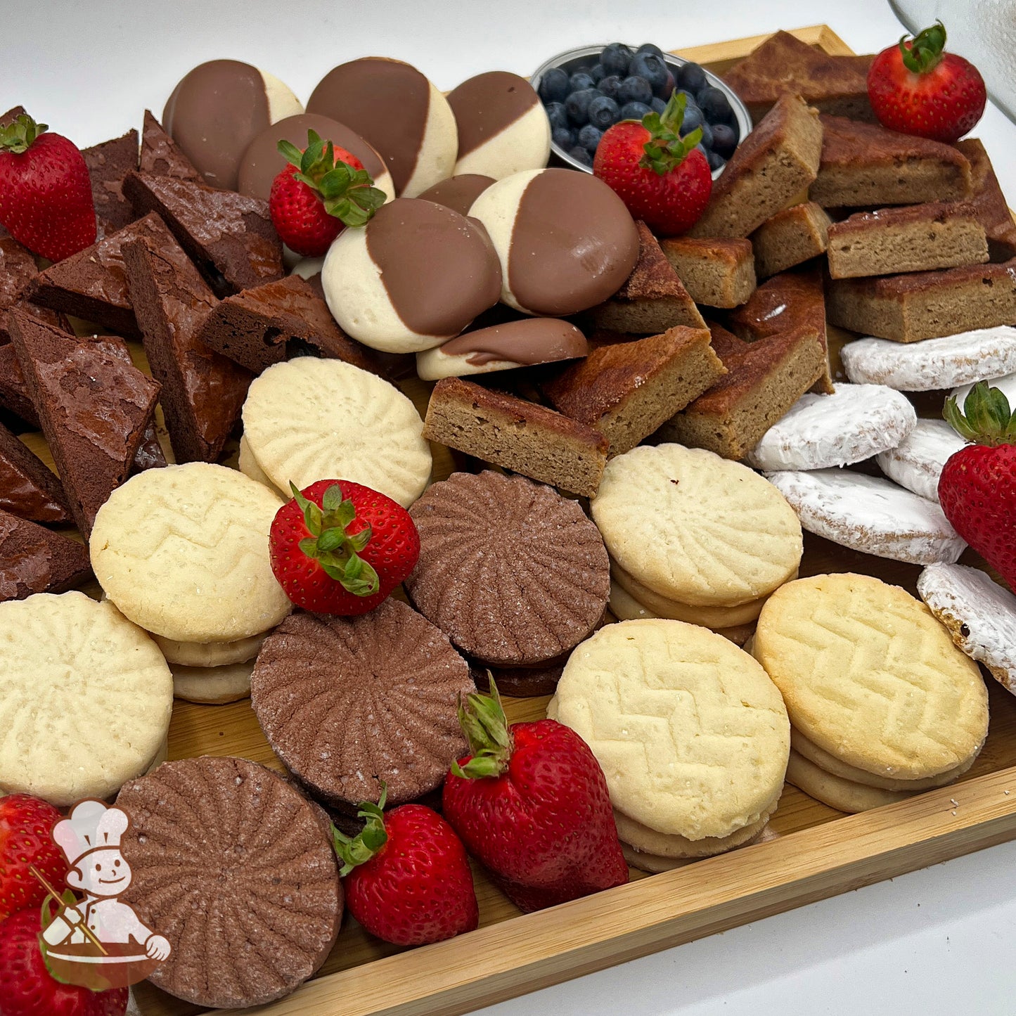 A rectangular bamboo tray carrying an assortment of cookies and brownies, fresh blueberries in a bowl, and strawberries.