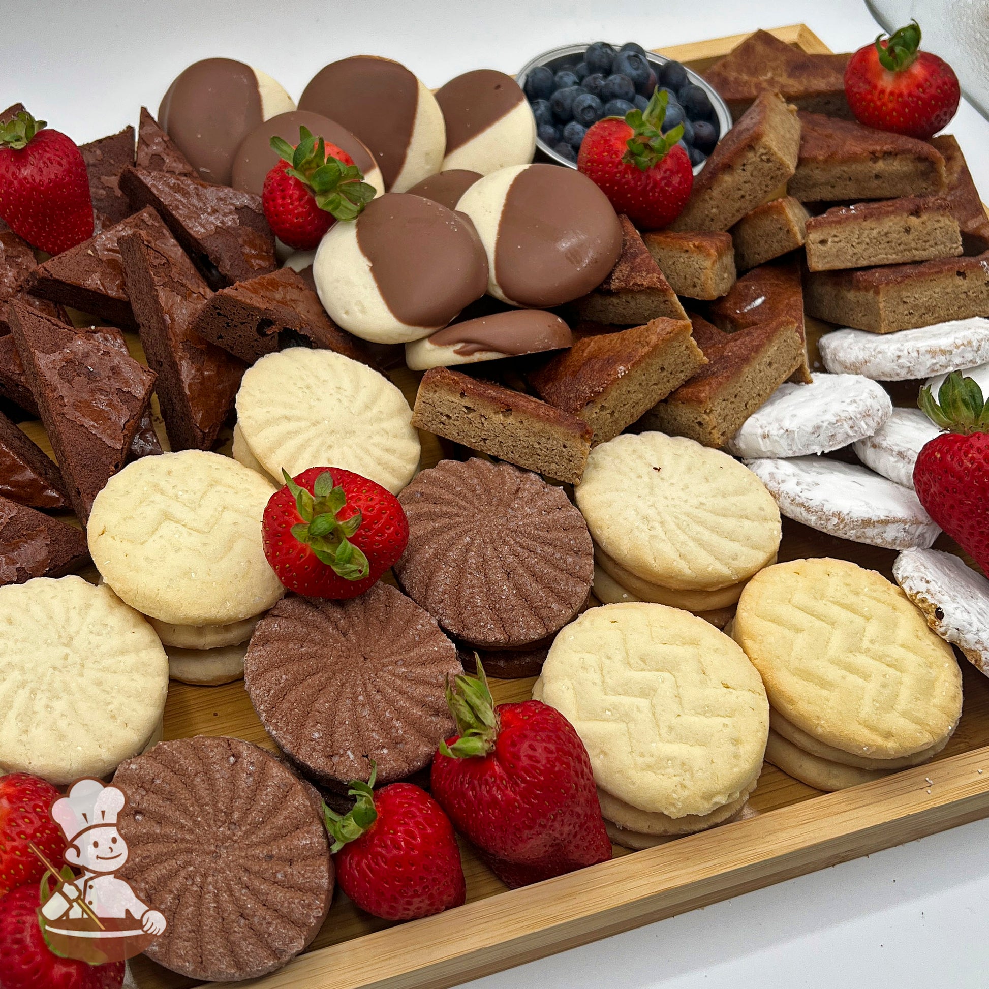 A rectangular bamboo tray carrying an assortment of cookies and brownies, fresh blueberries in a bowl, and strawberries.