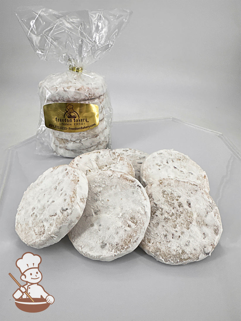 Six round shortbread cookies dusted with powdered sugar, arranged on a plate. A clear bag with more cookies is in the background.