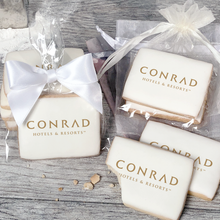 Load image into Gallery viewer, A stack of rectangular butter shortbread cookies with Conrad Hotels and Resorts logo printed directly on a white, light sugar icing. Some cookies are shown in clear packaging with a twist-tie ribbon bow or inside an organza bag.