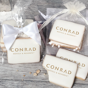 A stack of rectangular butter shortbread cookies with Conrad Hotels and Resorts logo printed directly on a white, light sugar icing. Some cookies are shown in clear packaging with a twist-tie ribbon bow or inside an organza bag.