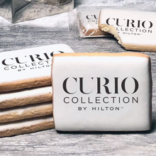 Load image into Gallery viewer, A stack of rectangle butter shortbread cookies with Curio Collection by Hilton logo printed directly on a white, lemon sugar icing.