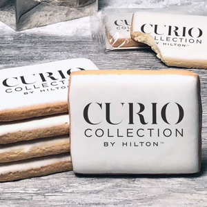 A stack of rectangle butter shortbread cookies with Curio Collection by Hilton logo printed directly on a white, lemon sugar icing.