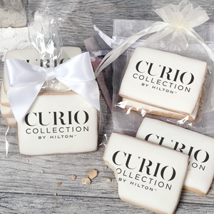 A stack of rectangular butter shortbread cookies with Curio Collection by Hilton logo printed directly on a white, light sugar icing. Some cookies are shown in clear packaging with a twist-tie ribbon bow or inside an organza bag.