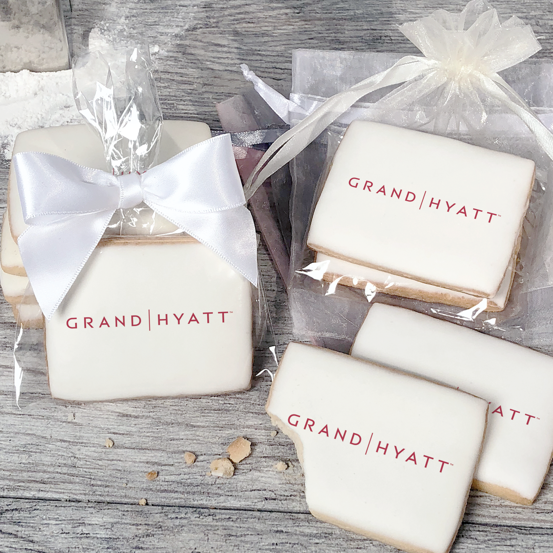 A stack of rectangular butter shortbread cookies with Grand Hyatt logo printed directly on a white, light sugar icing. Some cookies are shown in clear packaging with a twist-tie ribbon bow or inside an organza bag.
