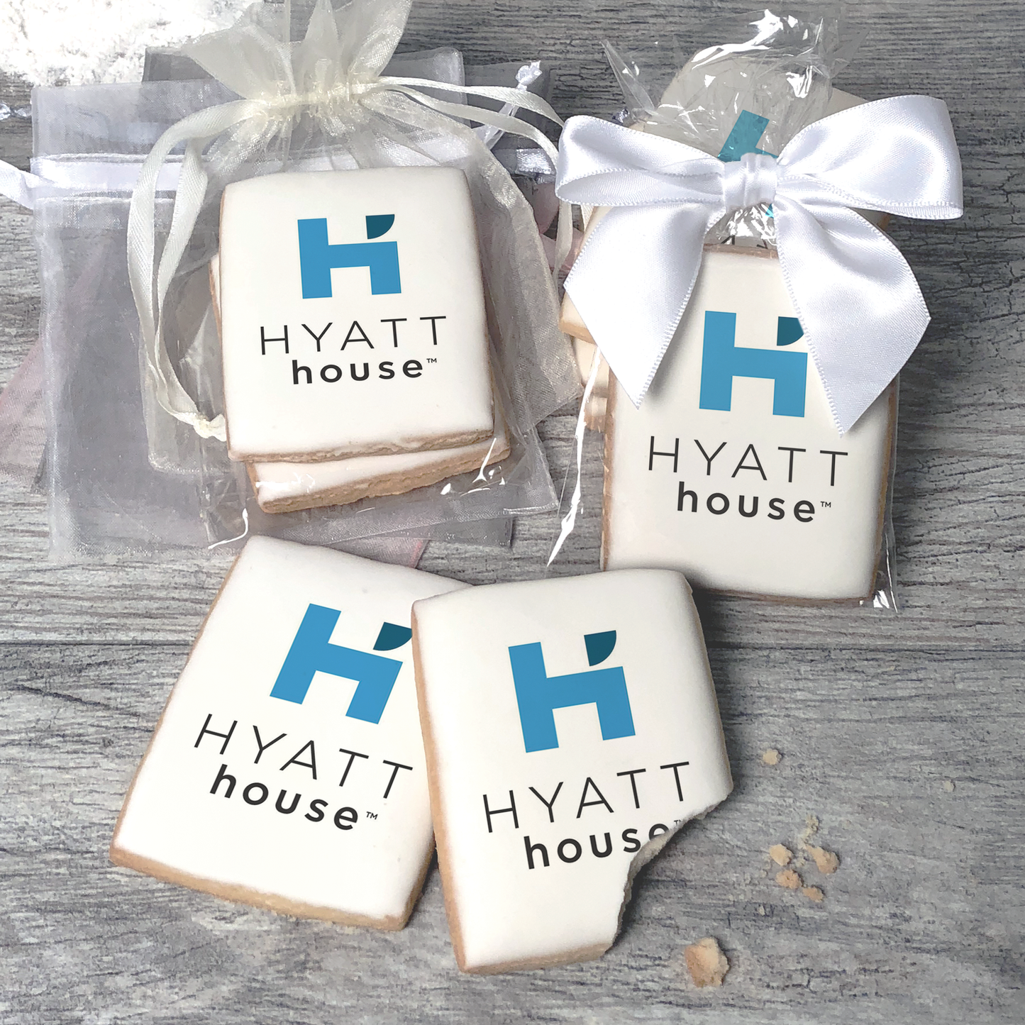 A stack of rectangular butter shortbread cookies with Hyatt House logo printed directly on a white, light sugar icing. Some cookies are shown in clear packaging with a twist-tie ribbon bow or inside an organza bag.