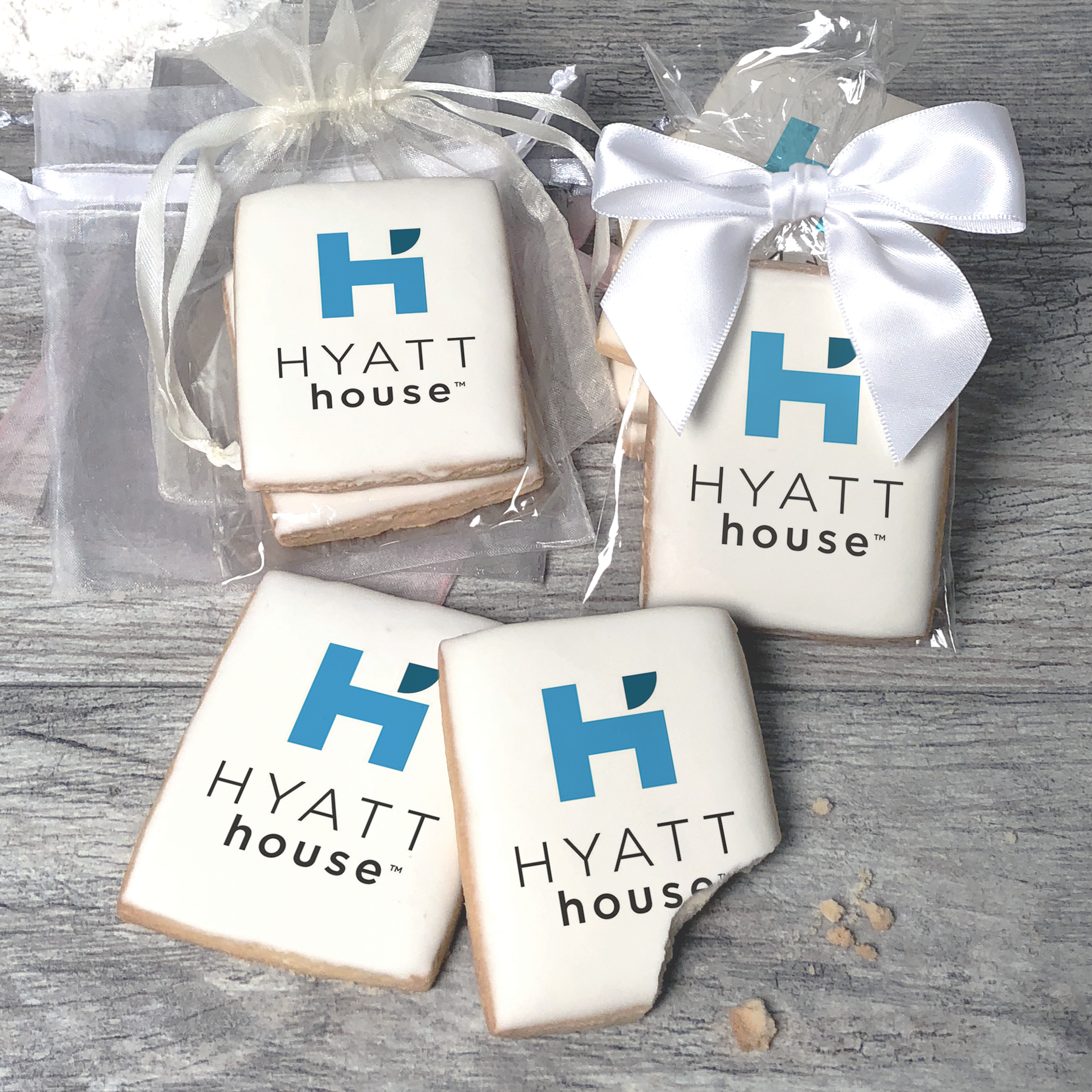 A stack of rectangular butter shortbread cookies with Hyatt House logo printed directly on a white, light sugar icing. Some cookies are shown in clear packaging with a twist-tie ribbon bow or inside an organza bag.