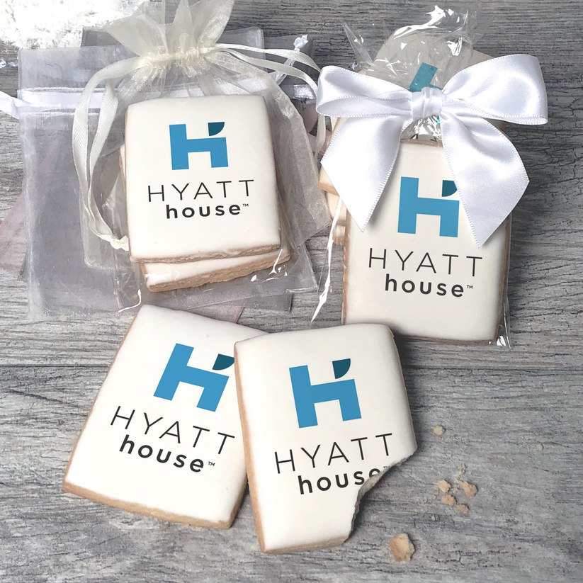 A stack of rectangular butter shortbread cookies with Hyatt House logo printed directly on a white, light sugar icing. Some cookies are shown in clear packaging with a twist-tie ribbon bow or inside an organza bag.
