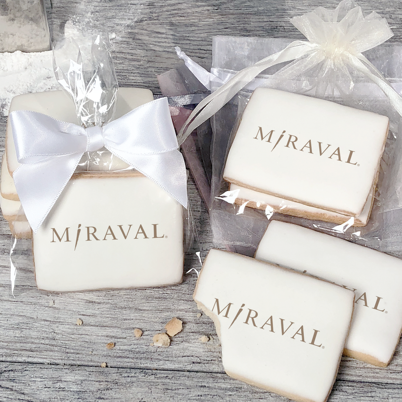 A stack of rectangular butter shortbread cookies with MiRaval logo printed directly on a white, light sugar icing. Some cookies are shown in clear packaging with a twist-tie ribbon bow or inside an organza bag.