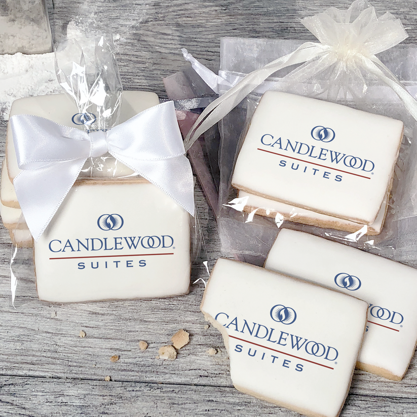 A stack of rectangular butter shortbread cookies with Candlewood Suites logo printed directly on a white, light sugar icing. Some cookies are shown in clear packaging with a twist-tie ribbon bow or inside an organza bag.