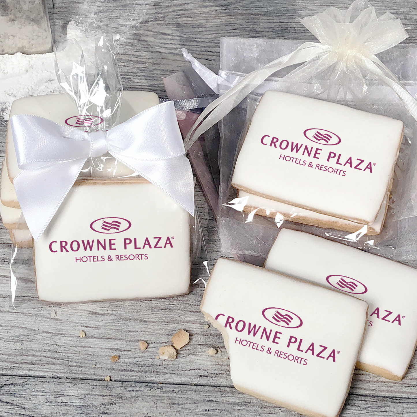 A stack of rectangular butter shortbread cookies with Crowne Plaza logo printed directly on a white, light sugar icing. Some cookies are shown in clear packaging with a twist-tie ribbon bow or inside an organza bag.