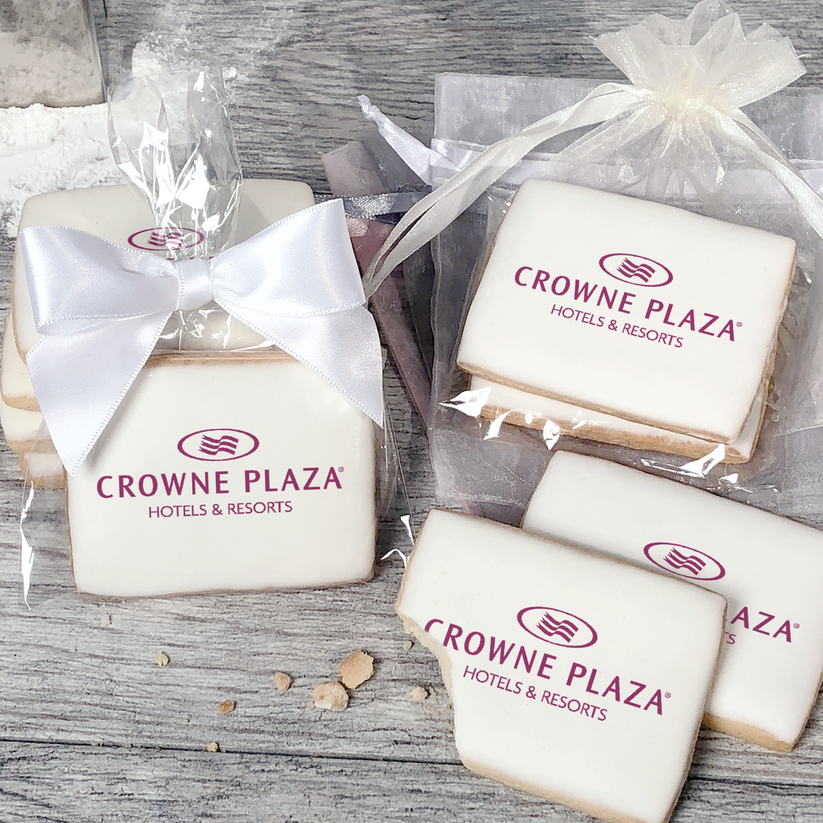 A stack of rectangular butter shortbread cookies with Crowne Plaza logo printed directly on a white, light sugar icing. Some cookies are shown in clear packaging with a twist-tie ribbon bow or inside an organza bag.