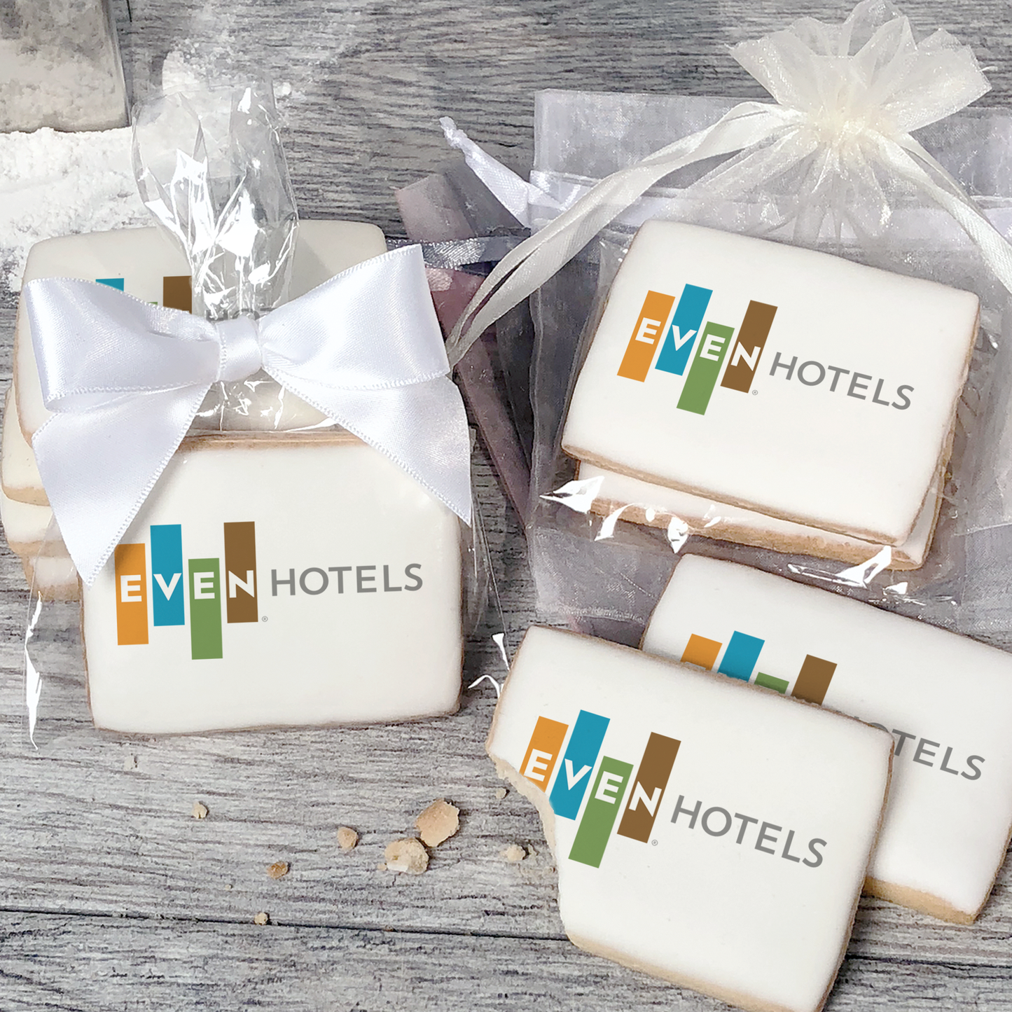 A stack of rectangular butter shortbread cookies with Even Hotels logo printed directly on a white, light sugar icing. Some cookies are shown in clear packaging with a twist-tie ribbon bow or inside an organza bag.