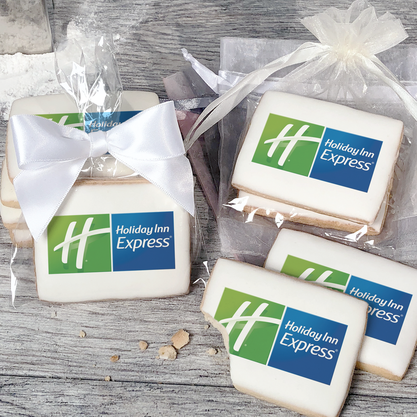A stack of rectangular butter shortbread cookies with Holiday Inn Express logo printed directly on a white, light sugar icing. Some cookies are shown in clear packaging with a twist-tie ribbon bow or inside an organza bag.