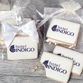 A stack of rectangular butter shortbread cookies with Hotel Indigo logo printed directly on a white, light sugar icing. Some cookies are shown in clear packaging with a twist-tie ribbon bow or inside an organza bag.
