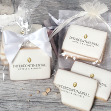 Load image into Gallery viewer, A stack of rectangular butter shortbread cookies with Intercontinental logo printed directly on a white, light sugar icing. Some cookies are shown in clear packaging with a twist-tie ribbon bow or inside an organza bag.
