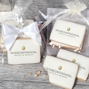 A stack of rectangular butter shortbread cookies with Intercontinental logo printed directly on a white, light sugar icing. Some cookies are shown in clear packaging with a twist-tie ribbon bow or inside an organza bag.